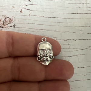 May include: Silver charm featuring a skull with a mustache. The skull has detailed eye sockets and a small hole at the top for attaching to a chain or bracelet.