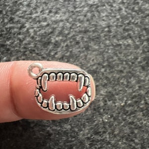 May include: Silver charm in the shape of a set of fangs. The charm has a loop at the top for attaching to a necklace or bracelet.