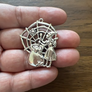 May include: Silver charm featuring a witch stirring a cauldron in a spiderweb. The witch is wearing a pointed hat and a long dress.