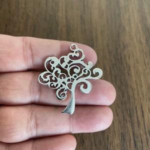 May include: A silver-colored tree charm with an intricate, openwork design. The charm features a detailed tree silhouette with a slender trunk and a rounded, decorative crown. It has a small loop at the top for attaching to jewelry.
