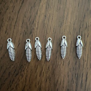 May include: Six silver corn on the cob charms. The charms are detailed with a textured surface and a loop at the top for attaching to jewelry or other crafts.