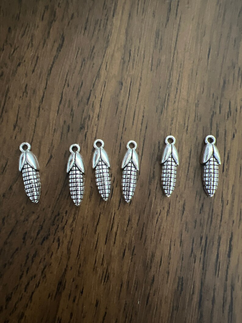 May include: Six silver corn on the cob charms. The charms are detailed with a textured surface and a loop at the top for attaching to jewelry or other crafts.