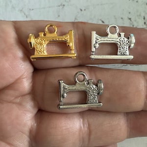 May include: Three sewing machine charms, one gold and two silver, with intricate details. The charms are on a hand.