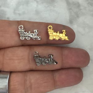 May include: Three silver train charms, one gold train charm. The charms are small and detailed, with a locomotive and a few train cars. The charms are on a hand, ready to be used for jewelry making.