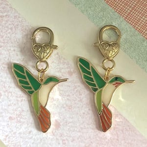 May include: Two gold-colored hummingbird charms with green, white, and orange enamel detailing. Each charm has a heart-shaped clasp. The hummingbird's wings are outstretched, and the design is detailed.