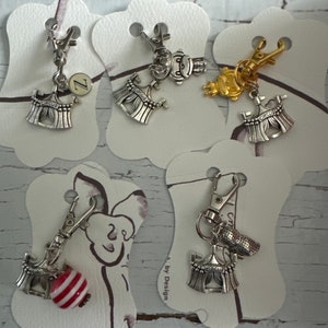 May include: Five silver charm keychains with a circus tent design. Each keychain has a different charm attached to the tent, including a letter Z, a small figure, a gold charm, and a red and white striped bead.