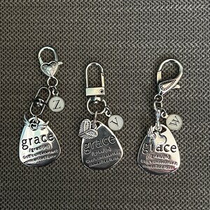 May include: Three silver keychains with a heart charm and a small charm with a letter on it. Each keychain has a silver oval charm with the word "grace" and a definition of the word. The charms are on a silver chain with a silver lobster clasp.