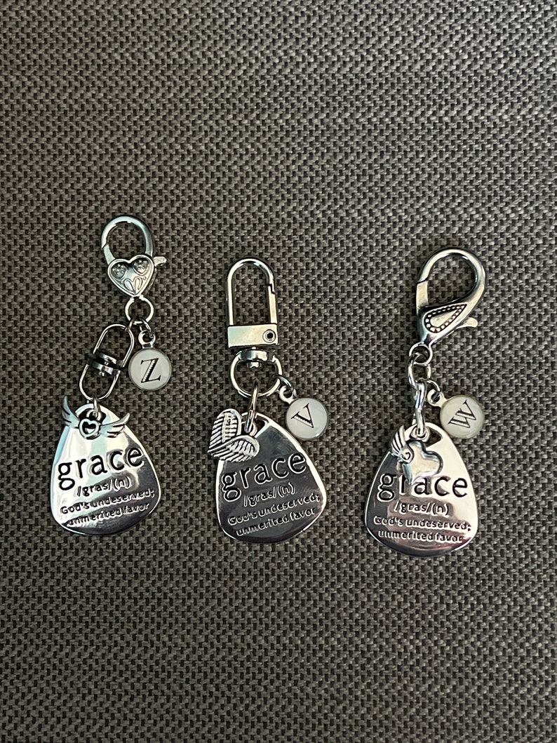 May include: Three silver keychains with a heart charm and a small charm with a letter on it. Each keychain has a silver oval charm with the word "grace" and a definition of the word. The charms are on a silver chain with a silver lobster clasp.