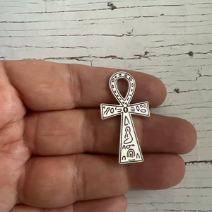 May include: Silver Ankh pendant with intricate details and hieroglyphics. The pendant is held in a hand.
