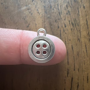 May include: A small, silver-colored button charm with four holes in a square pattern. The charm has a loop at the top for attaching to a necklace or bracelet. The button charm is a craft supply.