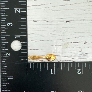 May include: A small gold-colored spoon charm with a loop for attaching to a necklace or bracelet. The charm is about 1 inch long and is shown next to a ruler.