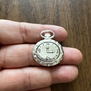 May include: Silver charm in the shape of a pocket watch with Roman numeral markings and a hand pointing to the number 3.