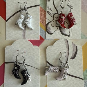 May include: Four pairs of dangle earrings featuring miniature boot charms. The boots are in white, red, black, and pink with silver accents and rhinestones.