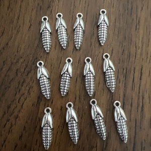 May include: A collection of 14 silver-toned metal charms shaped like ears of corn. The charms have a detailed design and are perfect for adding a touch of rustic charm to jewelry or crafts.