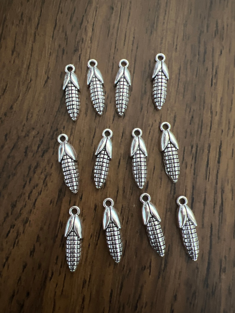 May include: A collection of 14 silver-toned metal charms shaped like ears of corn. The charms have a detailed design and are perfect for adding a touch of rustic charm to jewelry or crafts.