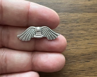May include: Silver charm in the shape of a stylized angel with wings outstretched. The angel has a halo and a small body.