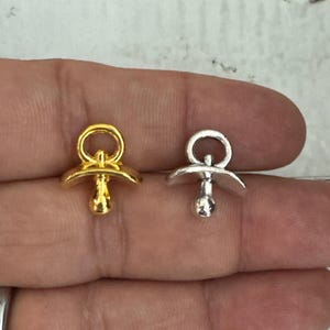 May include: Two pacifier charms, one gold and one silver, are shown on a hand. The charms are small and have a loop at the top for attaching to a necklace or bracelet.