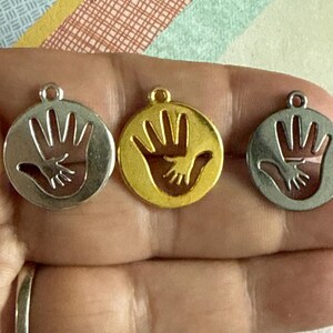 May include: Three silver and gold charms with a cutout design of two hands holding each other. The charms are round and have a smooth finish.