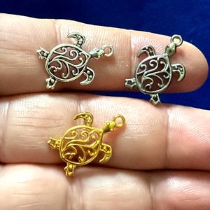 May include: Three metal turtle charms featuring intricate filigree cutouts on their shells. Two charms are silver-toned, and one charm is gold-toned. Each turtle charm includes a small circular loop at the head for attachment to jewelry or crafts.