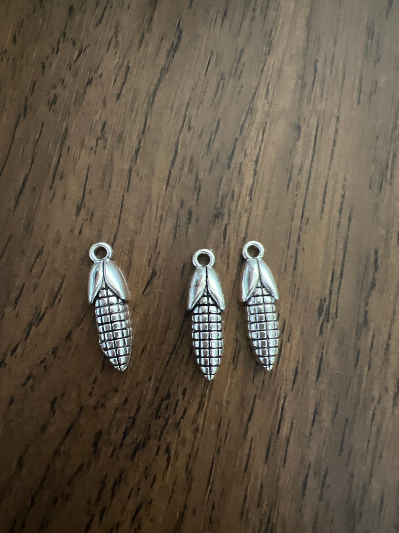 May include: Three silver charm pendants shaped like ears of corn. The charms are detailed with a textured surface to resemble corn kernels.