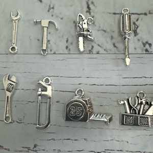 May include: A set of nine silver-toned metal charms shaped like various tools, including a wrench, hammer, saw, screwdriver, and a tape measure with the number 25 printed on it. The charms are arranged in two rows on a white wooden surface.