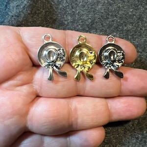 May include: Three metal hat charms in silver and gold tones. Each charm features a detailed hat design with a bow and decorative elements. The charms are small, suitable for jewelry making or crafting projects.