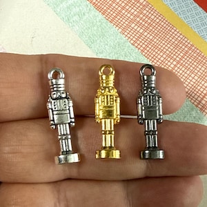 May include: Three nutcracker charms in silver, gold, and silver. The charms are detailed with a soldier's uniform and a hat. The charms are hanging from a hand.