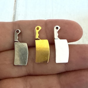 May include: Three miniature cleaver charms in silver, gold, and silver tones. Each charm features a curved blade and a handle with a small, dark accent. The charms are displayed against a neutral background.
