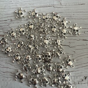 K&ouml;nnte beinhalten: Ein Haufen silberner Totenkopf-Anh&auml;nger mit Kreuzknochen. Die Anh&auml;nger haben oben eine Schlaufe zum Anbringen an Schmuck oder anderen Bastelarbeiten.