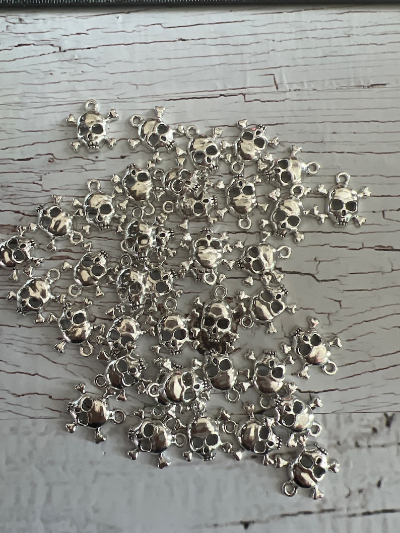 K&ouml;nnte beinhalten: Ein Haufen silberner Totenkopf-Anh&auml;nger mit Kreuzknochen. Die Anh&auml;nger haben oben eine Schlaufe zum Anbringen an Schmuck oder anderen Bastelarbeiten.