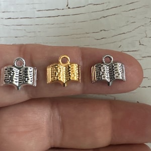 May include: Three silver and gold book charms, each with a detailed open book design. The charms are on a finger, showing their size.