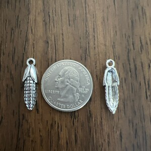 May include: Two silver charms, one shaped like an ear of corn and the other shaped like a feather, are shown next to a US quarter dollar coin for size comparison.