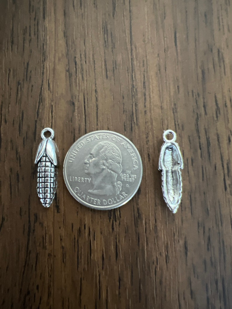 May include: Two silver charms, one shaped like an ear of corn and the other shaped like a feather, are shown next to a US quarter dollar coin for size comparison.