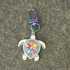May include: A silver-toned turtle keychain with a rainbow-colored flower design in the center. The turtle has a turquoise border and a multi-colored clasp. The keychain is designed to attach to keys or a bag.