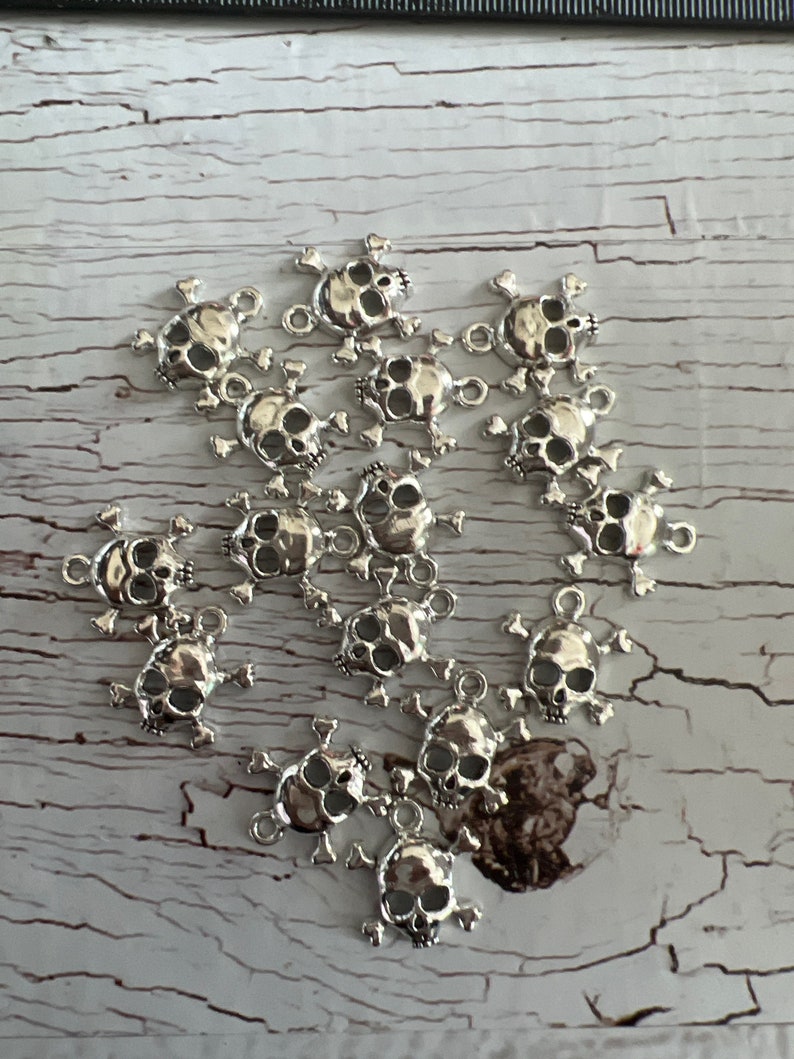 K&ouml;nnte beinhalten: Eine Sammlung silberner Totenkopf-Anh&auml;nger mit Kreuzknochen. Die Anh&auml;nger sind detailliert und perfekt, um Schmuck oder Bastelarbeiten einen Hauch von Gothic-Stil zu verleihen.