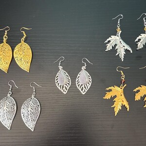 May include: A collection of leaf-shaped earrings in gold and silver tones. The earrings feature intricate cut-out designs and are displayed on a dark surface. Some earrings have decorative beads.