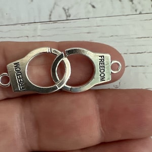 May include: Silver-toned metal charm in the shape of handcuffs with the word "FREEDOM" engraved on each side.