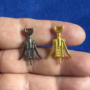 May include: Two small metal corkscrew charms, one in a dark gunmetal grey finish and the other in a bright gold tone. Each charm features a central cylindrical body, two side arms, a top handle, and a spiral worm at the bottom base.