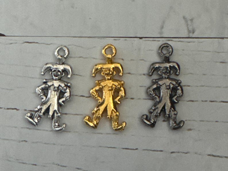 K&ouml;nnte beinhalten: Drei Jester-Anh&auml;nger, einer silber, einer gold und einer schwarz. Jeder Anh&auml;nger ist eine kleine, detaillierte Figur eines Jesters mit Hut und Gl&ouml;ckchen.