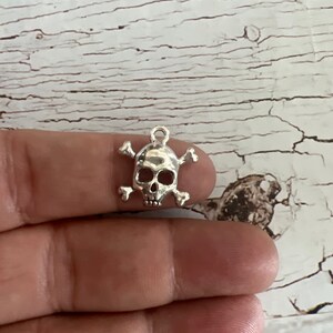 K&ouml;nnte beinhalten: Silberner Totenkopf- und Knochen-Anh&auml;nger. Der Totenkopf ist detailliert und die Knochen sind einfach.
