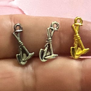 May include: Three small metal charms shaped like a shovel and a pickaxe crossed together. Two of the charms have a silver-toned finish, while the third charm on the right has a bright gold-toned finish. Each charm features a small loop at the top.