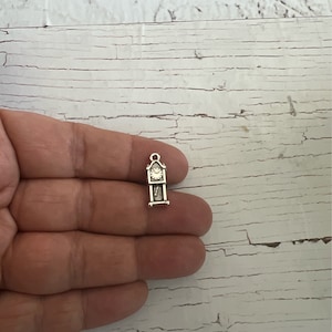 May include: Silver charm in the shape of a grandfather clock. The clock face has black Roman numeral markings and black hands.