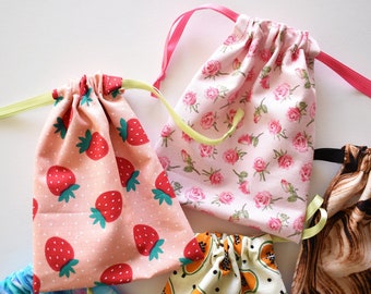 May include: A collection of drawstring bags in various patterns. One bag features red strawberries on a pink background, another has pink roses, and a third displays papaya slices. Each bag has a contrasting ribbon drawstring.