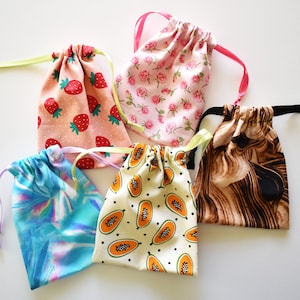 Könnte beinhalten: Fünf kleine Kordelzugbeutel mit verschiedenen Mustern. Eine Tasche zeigt Erdbeeren auf rosa Grund, eine andere rosa Rosen und eine dritte zeigt Papayastücke. Die restlichen Taschen haben ein blaues Batikmuster und ein braunes abstraktes Design.