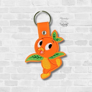 May include: Orange bird keychain with a metal ring and orange leather strap. The bird has a cheerful expression with white eyes, a yellow beak, and green leaf accents. The keychain is against a white brick wall.