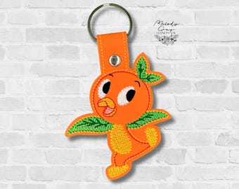 May include: Orange bird keychain with a metal ring and orange leather strap. The bird has a cheerful expression with white eyes, a yellow beak, and green leaf accents. The keychain is against a white brick wall.