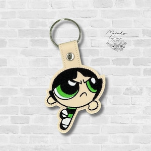 May include: Embroidered keychain featuring Buttercup from The Powerpuff Girls. The keychain has a cream-colored leather strap and a silver key ring. Buttercup is depicted with black hair, green eyes, and a determined expression. The design is outlined in black and green.
