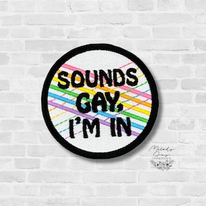 May include: Embroidered patch with the text "SOUNDS GAY, I'M IN" in black, set against a white background with colorful diagonal stripes. The patch has a black border and is likely for clothing or accessories.