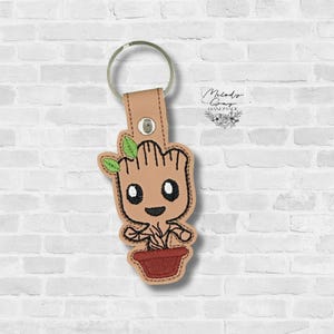 May include: A tan and brown embroidered keychain featuring a cartoon character resembling a tree with a smiling face and green leaves. The keychain has a metal ring and a leather strap. The character is in a brown pot. The background is a white brick wall.