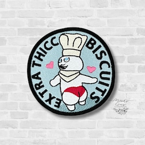 May include: Embroidered patch featuring a cartoon character in a chef's hat and red shorts, with the text "EXTRA THICC BISCUITS" in a circular design. Two pink hearts are on either side of the character. The patch has a black border.